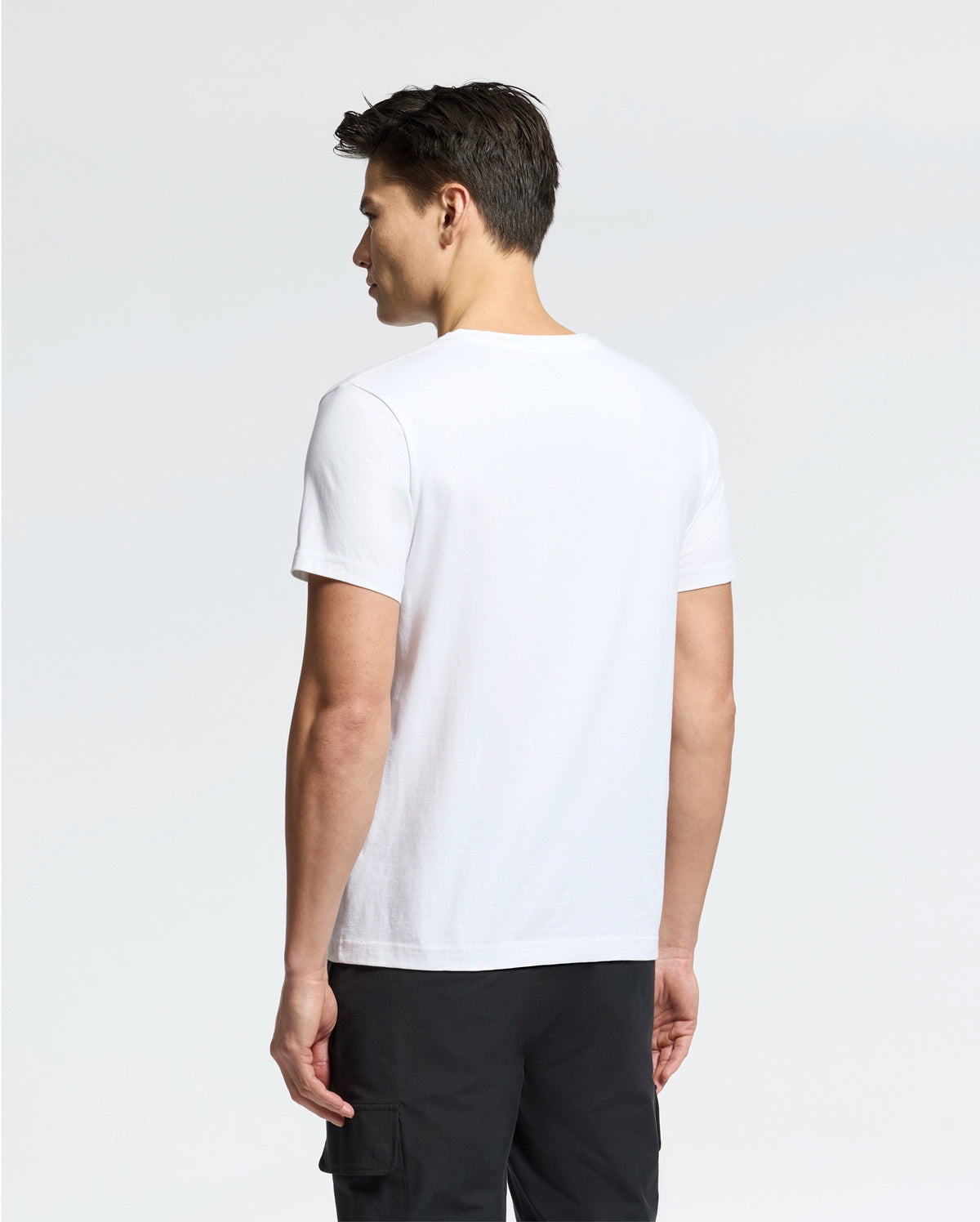 A person with short dark hair stands with their back to the camera, wearing a Psycho Bunny MENS CLASSIC V NECK TEE (B6U100ARPC) in plain white, made of 100% Pima cotton, and black pants. The background is neutral light gray.