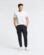 A person stands against a light gray background wearing a Psycho Bunny Men's Classic V Neck Tee (B6U100ARPC) made of 100% Pima cotton, paired with black cargo pants and white sneakers, hands in pockets, with a relaxed expression.