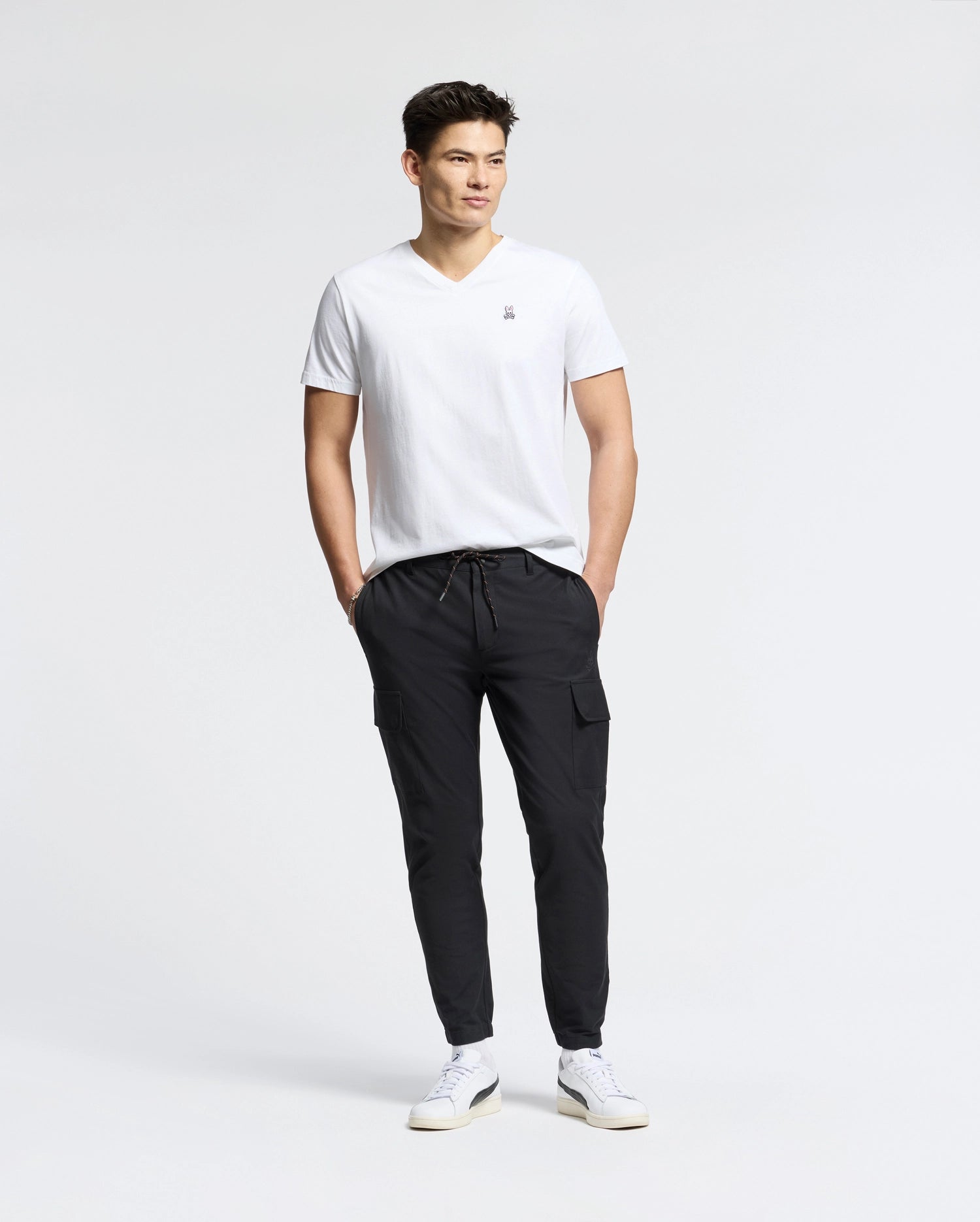 A person stands against a light gray background wearing a Psycho Bunny Men's Classic V Neck Tee (B6U100ARPC) made of 100% Pima cotton, paired with black cargo pants and white sneakers, hands in pockets, with a relaxed expression.