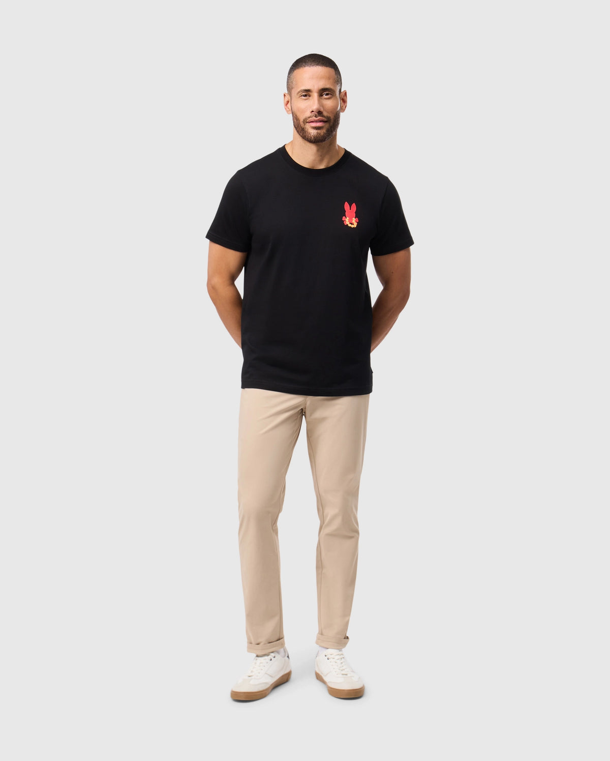 A man with short hair and a beard is wearing the Psycho Bunny MENS HAWAII CITY TEE in black Pima cotton, featuring a red design on the chest, along with beige pants and white sneakers. He stands against a simple light gray background, with his hands positioned behind his back.