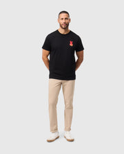 A man with short hair and a beard is wearing the Psycho Bunny MENS HAWAII CITY TEE in black Pima cotton, featuring a red design on the chest, along with beige pants and white sneakers. He stands against a simple light gray background, with his hands positioned behind his back.