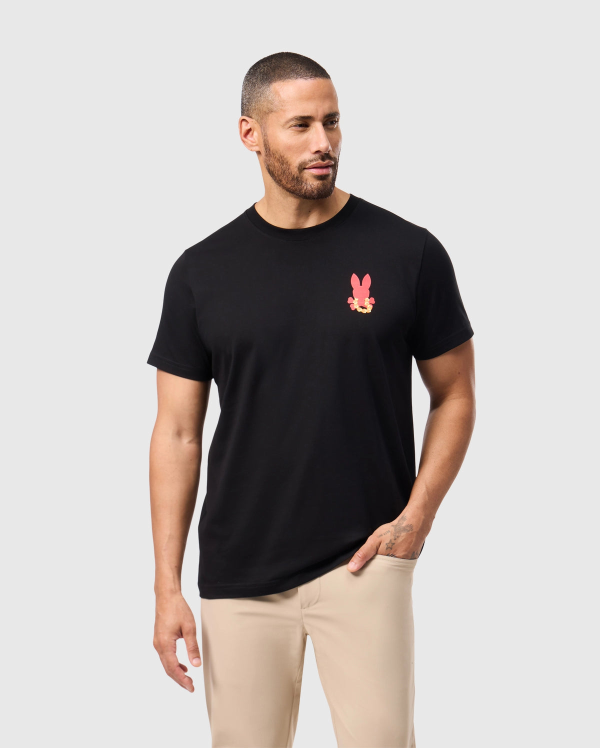A man with short hair and a beard is wearing the black Psycho Bunny MENS HAWAII CITY TEE - B6U169Y1PC, which features a pink rabbit graphic on the chest. This tee is made from soft Pima cotton. He stands against a light gray background, looking to his right, and pairs it with beige pants for a relaxed look.