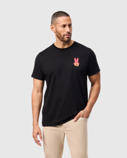 A man with short hair and a beard is wearing the black Psycho Bunny MENS HAWAII CITY TEE - B6U169Y1PC, which features a pink rabbit graphic on the chest. This tee is made from soft Pima cotton. He stands against a light gray background, looking to his right, and pairs it with beige pants for a relaxed look.