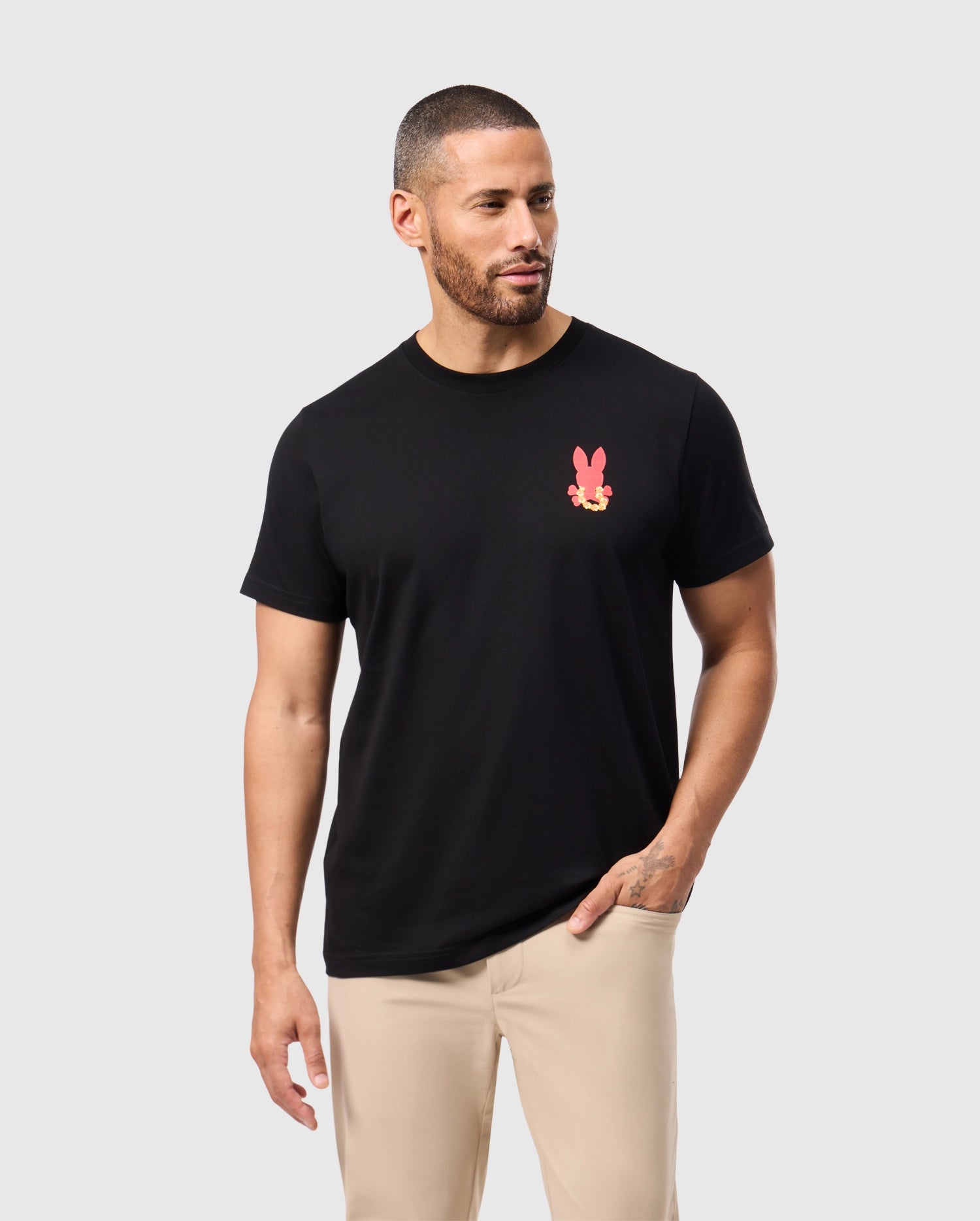 A man with short hair and a beard is wearing the black Psycho Bunny MENS HAWAII CITY TEE - B6U169Y1PC, which features a pink rabbit graphic on the chest. This tee is made from soft Pima cotton. He stands against a light gray background, looking to his right, and pairs it with beige pants for a relaxed look.