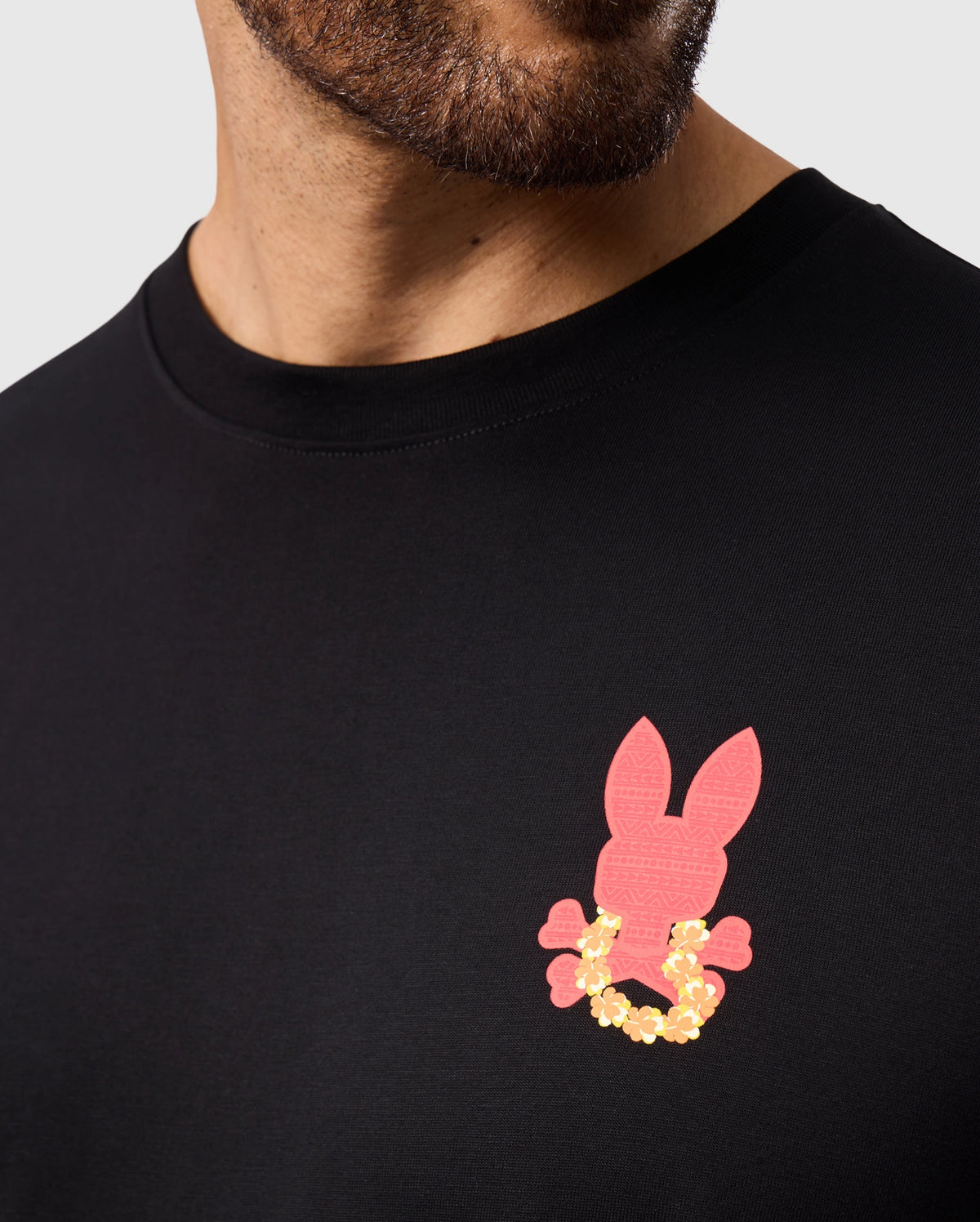 A close-up captures someone wearing the Psycho Bunny MENS HAWAII CITY TEE - B6U169Y1PC. This black tee, made from soft Pima cotton, showcases a red rabbit head graphic and a yellow floral design on the chest while partially obscuring the person's face to maintain their anonymity.
