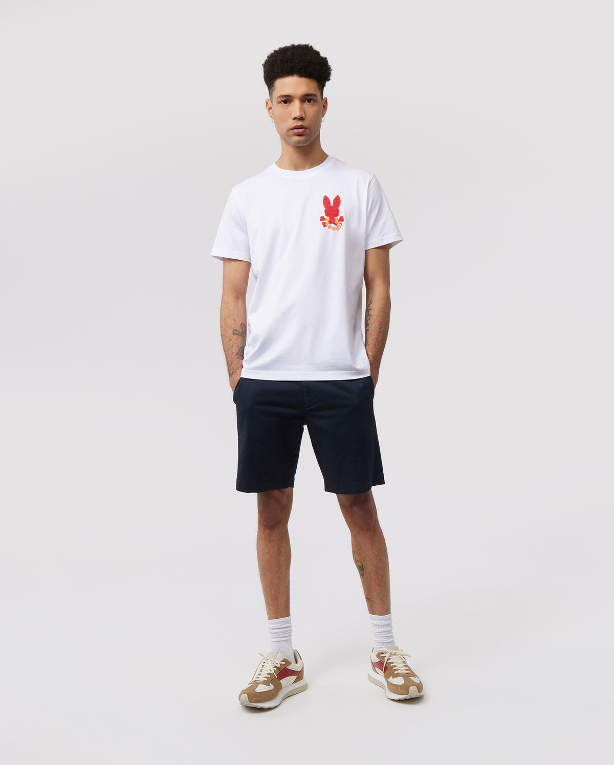 A young man stands against a plain white background. He is wearing a white Psycho Bunny MENS HAWAII CITY TEE - B6U169Y1PC with a red rabbit graphic on the left chest, navy blue shorts, white socks, and beige sneakers with red accents. He has short curly hair and his hands are by his sides.