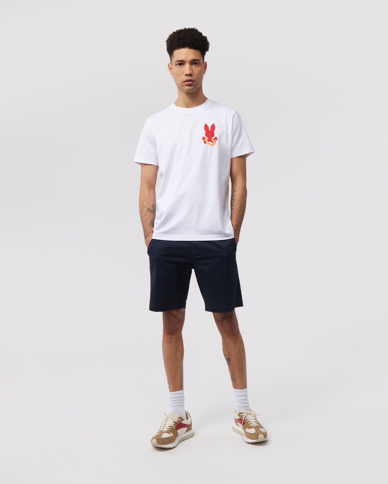 A young man stands against a plain white background. He is wearing a white Psycho Bunny MENS HAWAII CITY TEE - B6U169Y1PC with a red rabbit graphic on the left chest, navy blue shorts, white socks, and beige sneakers with red accents. He has short curly hair and his hands are by his sides.