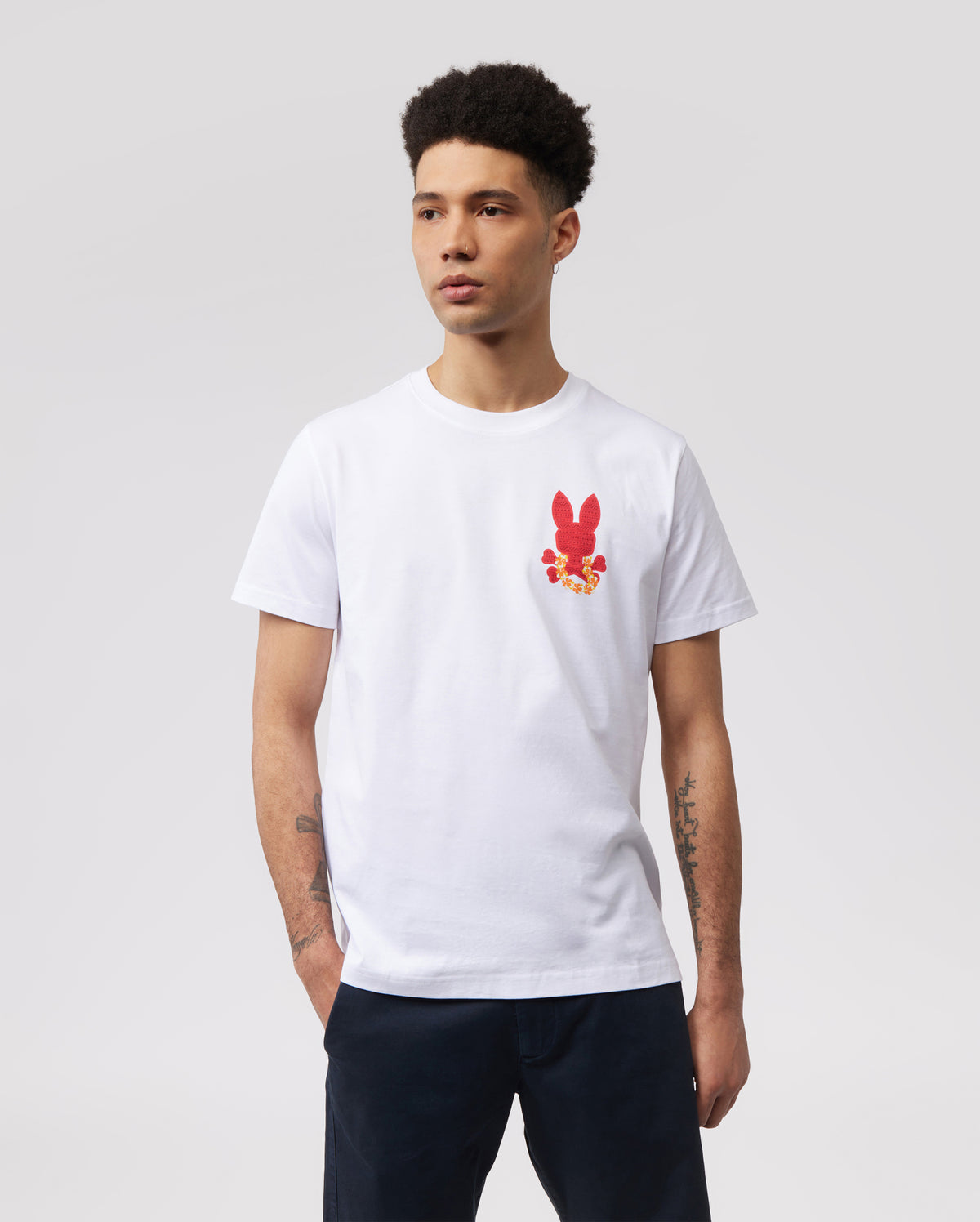 A person with short curly hair is wearing a white MENS HAWAII CITY TEE - B6U169Y1PC by Psycho Bunny made of soft Pima cotton jersey, featuring a small red bunny graphic pattern on the left side of the chest. Their hands are in their pockets, and they are standing against a plain, light gray background.