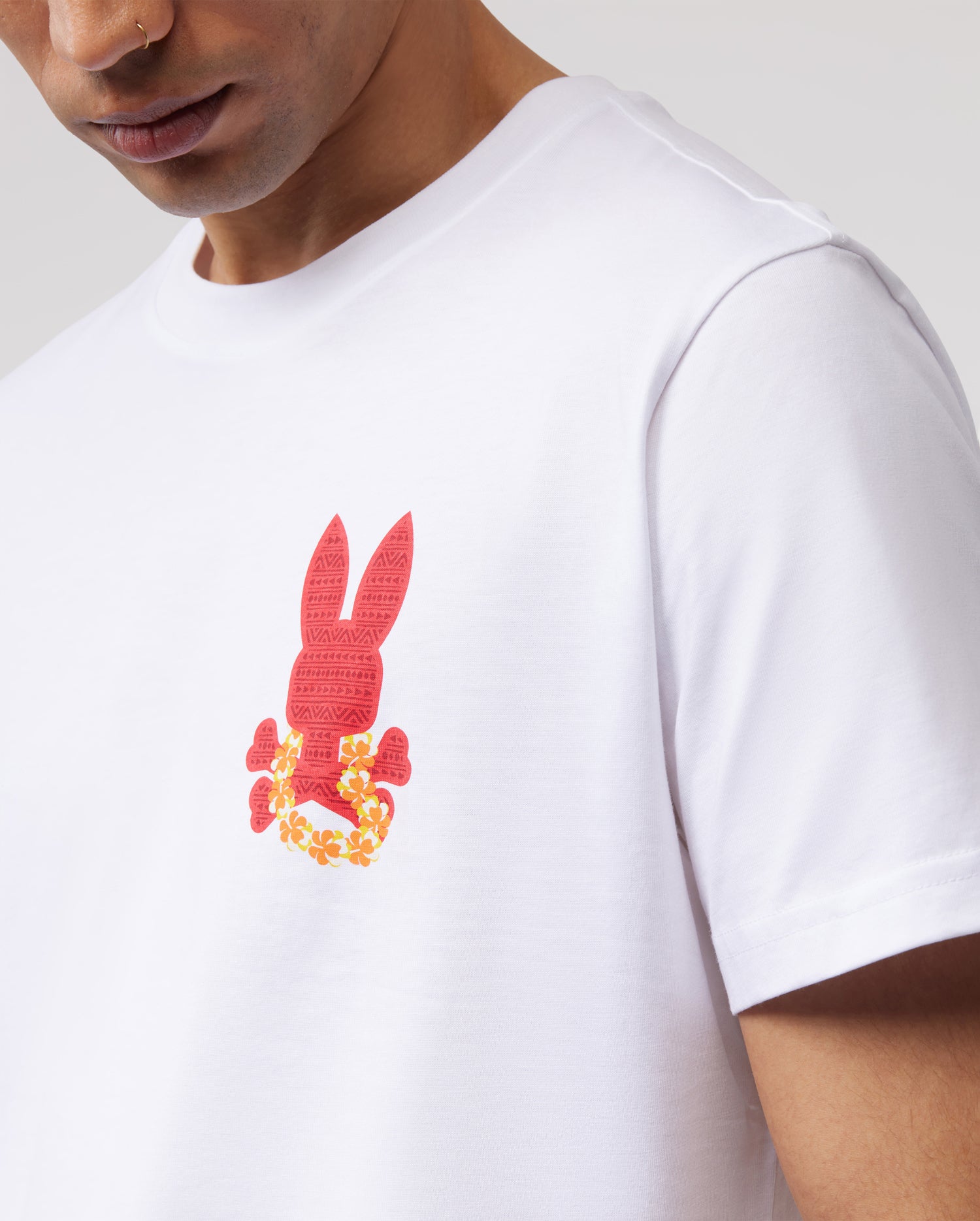 A person wearing a white Psycho Bunny MENS HAWAII CITY TEE - B6U169Y1PC featuring a red design of a bunny with vibrant orange-yellow flowers at the bottom. The individual has a nose ring and is shown from the shoulders up with their face partially out of frame.