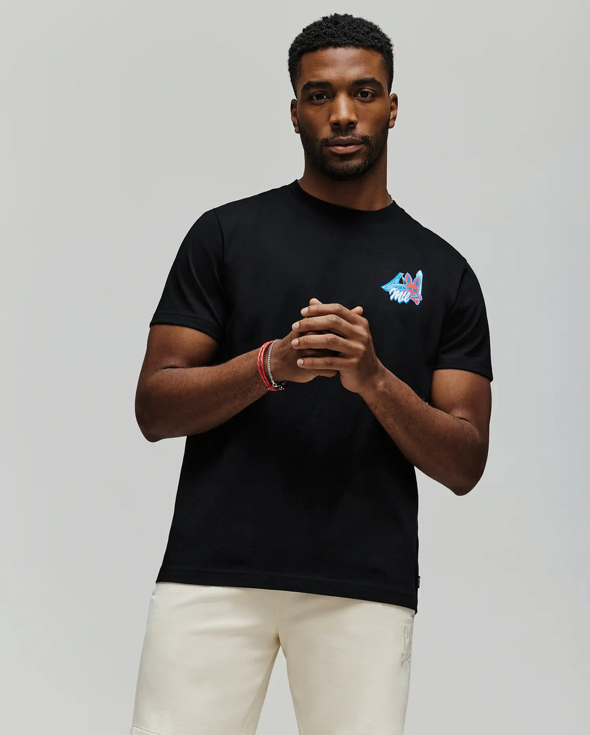 A man with short hair and a beard wears the Psycho Bunny MENS MONTREAL TEE (B6U381F200), featuring a small colorful chest graphic, paired with light shorts, standing against a plain gray background with his hands loosely clasped.