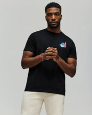 A man with short hair and a beard wears the Psycho Bunny MENS MONTREAL TEE (B6U381F200), featuring a small colorful chest graphic, paired with light shorts, standing against a plain gray background with his hands loosely clasped.
