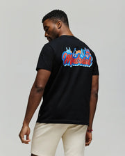 A man stands facing away, wearing a Psycho Bunny MENS MONTREAL TEE (B6U381F200) in black, featuring colorful “Montreal” text and city skyline graphics on soft Pima cotton, paired with light shorts against a plain background.