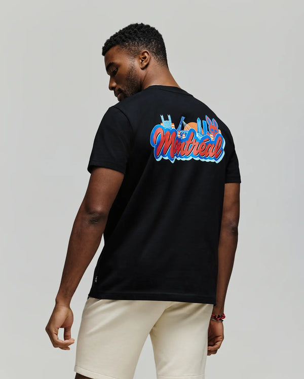 A man stands facing away, wearing a Psycho Bunny MENS MONTREAL TEE (B6U381F200) in black, featuring colorful “Montreal” text and city skyline graphics on soft Pima cotton, paired with light shorts against a plain background.