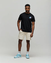 A man poses against a plain background wearing the Psycho Bunny MENS MONTREAL TEE - B6U381F200 in black, paired with beige shorts, white socks, and light blue sneakers.