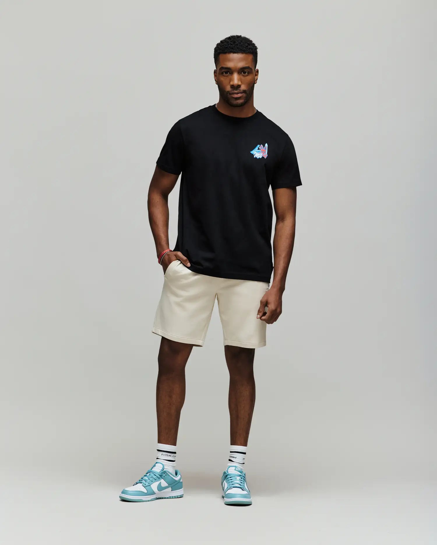 A man poses against a plain background wearing the Psycho Bunny MENS MONTREAL TEE - B6U381F200 in black, paired with beige shorts, white socks, and light blue sneakers.