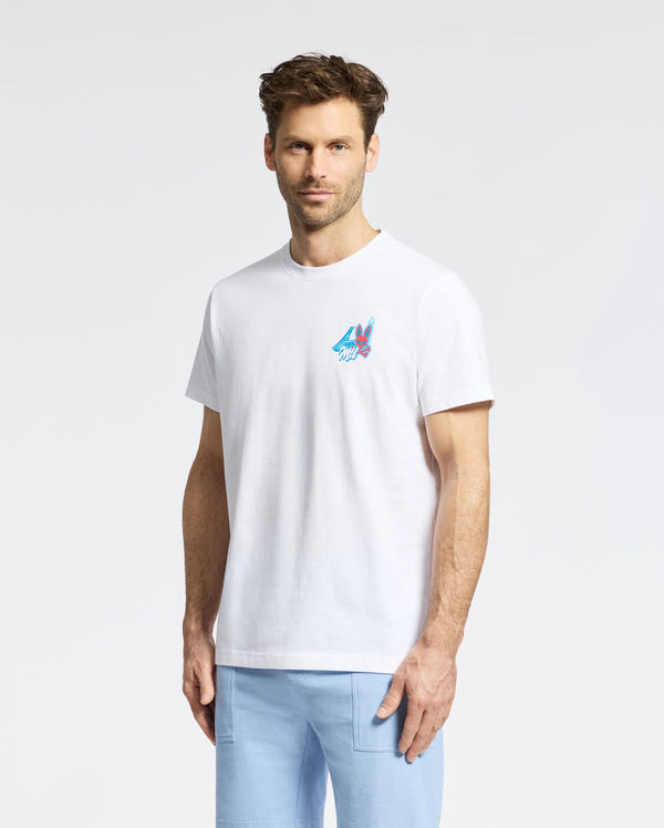 A man with short brown hair and a beard wears the Psycho Bunny MENS MONTREAL TEE (B6U381F200), a white Pima cotton T-shirt with a small colorful chest graphic, paired with light blue pants, standing against a plain background.