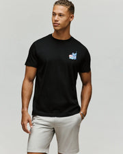 A man poses against a plain background wearing the Psycho Bunny MENS TORONTO TEE - B6U383F200 in black, featuring a colorful hometown hero graphic on the left chest, paired with light gray shorts and a relaxed expression.