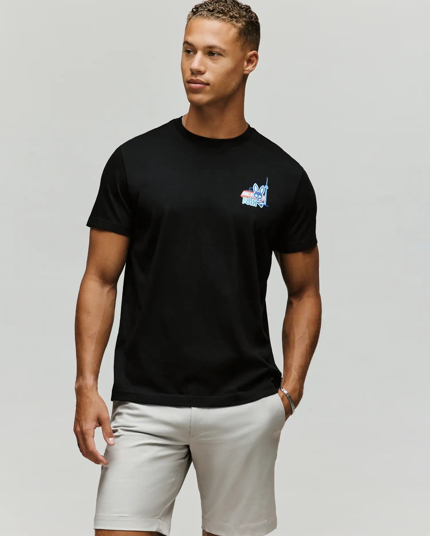 A man poses against a plain background wearing the Psycho Bunny MENS TORONTO TEE - B6U383F200 in black, featuring a colorful hometown hero graphic on the left chest, paired with light gray shorts and a relaxed expression.