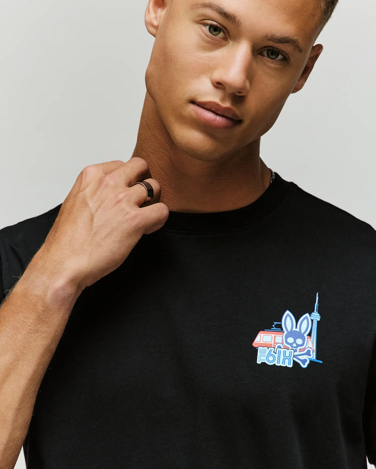 A young man with light skin and short hair wears the Psycho Bunny MENS TORONTO TEE (B6U383F200), a black Pima cotton shirt featuring a small hometown hero graphic of a bunny, red van, and CN Tower. He looks at the camera adjusting his collar.