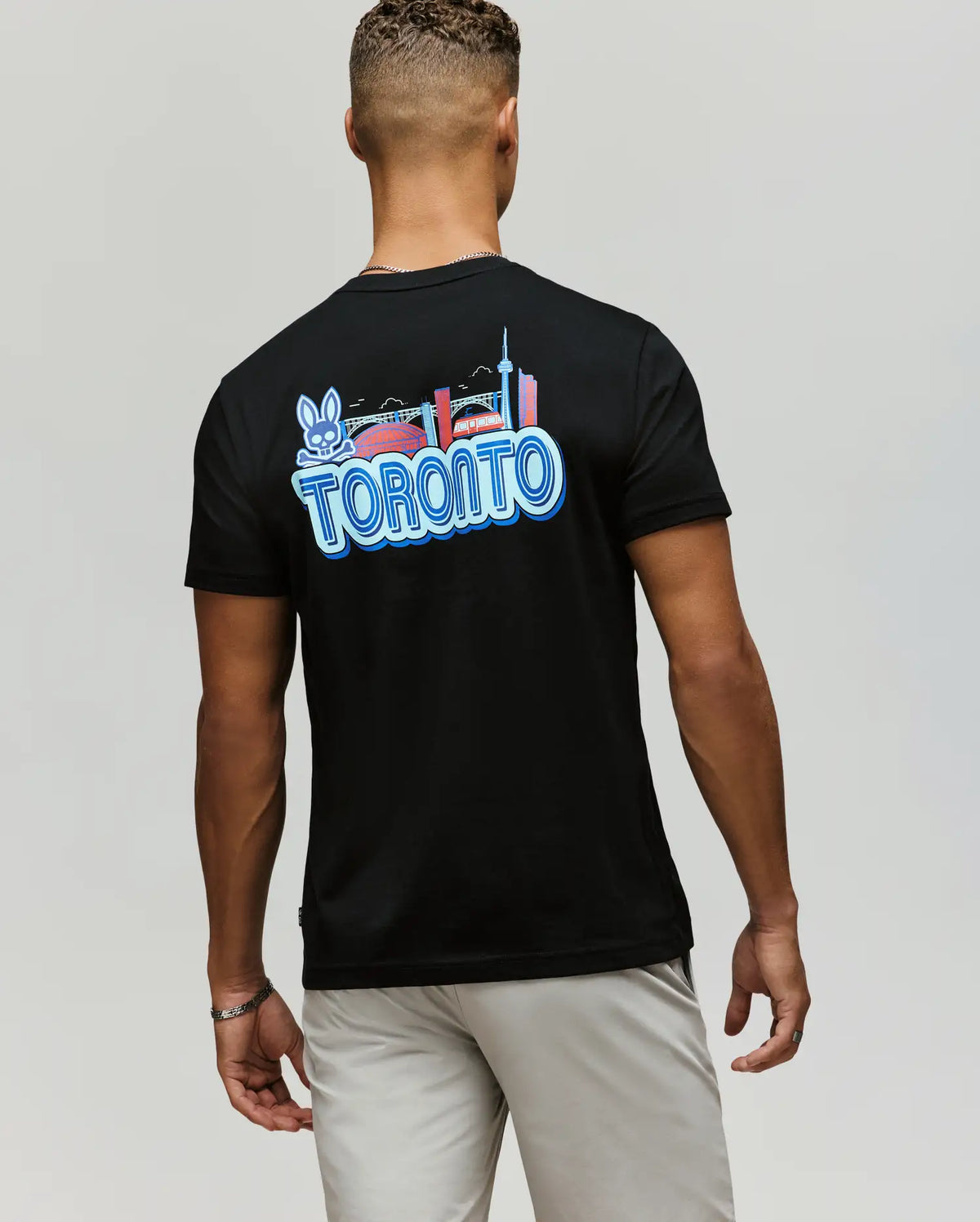 A man wears the Psycho Bunny MENS TORONTO TEE (B6U383F200), featuring a colorful hometown hero graphic with city landmarks, a bridge, and the bunny logo on the back. Made of soft Pima cotton, he pairs it with light shorts, standing with his back to the camera.