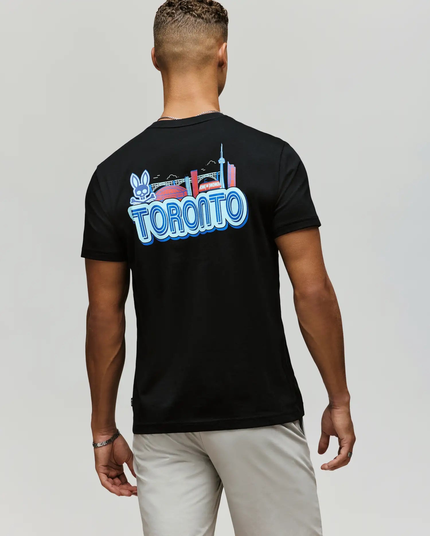 A man wears the Psycho Bunny MENS TORONTO TEE (B6U383F200), featuring a colorful hometown hero graphic with city landmarks, a bridge, and the bunny logo on the back. Made of soft Pima cotton, he pairs it with light shorts, standing with his back to the camera.
