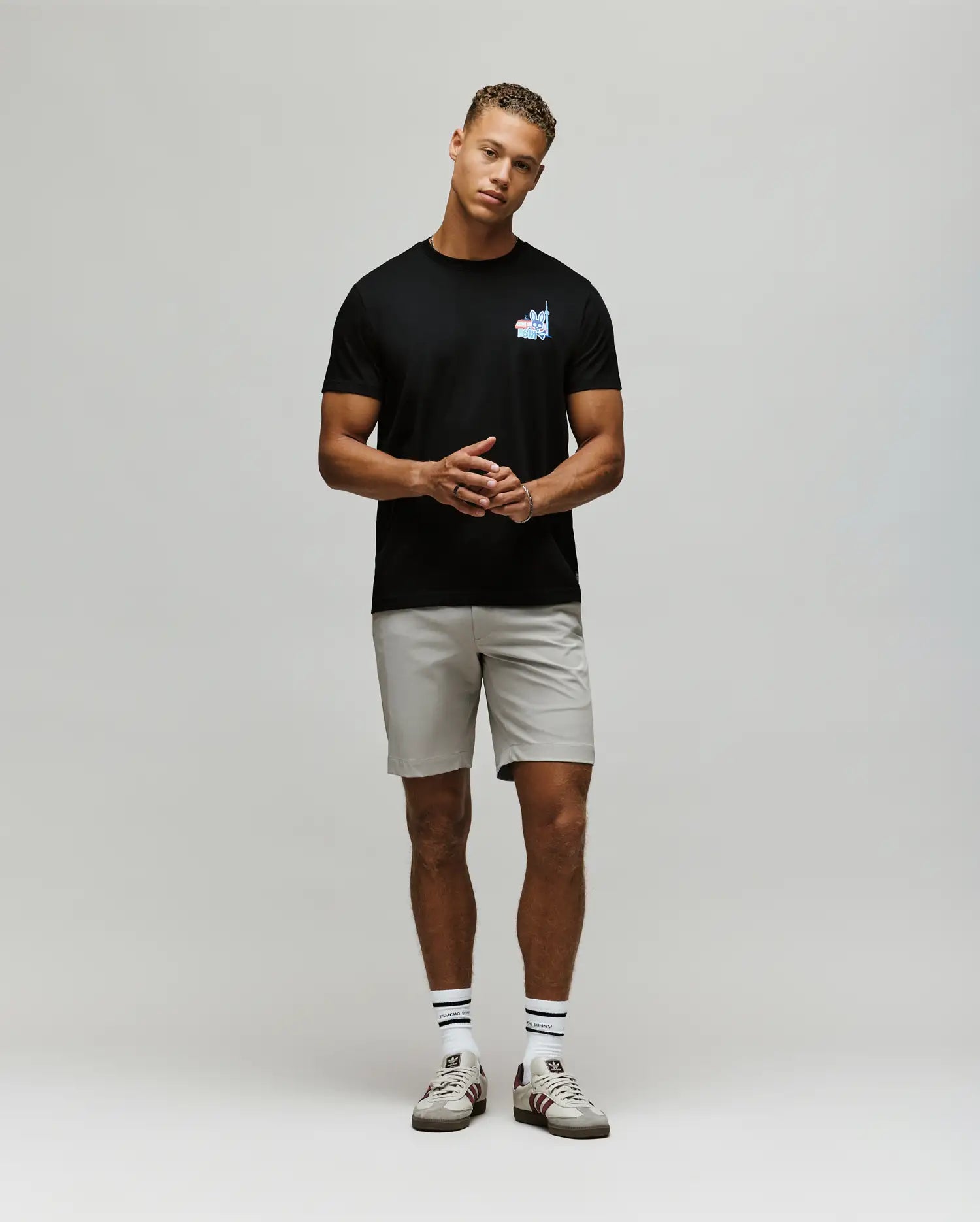 A man stands against a plain background wearing the Psycho Bunny MENS TORONTO TEE - B6U383F200 in black Pima cotton, paired with light gray shorts, white socks with black stripes, and white sneakers featuring red accents. His hands are loosely clasped.