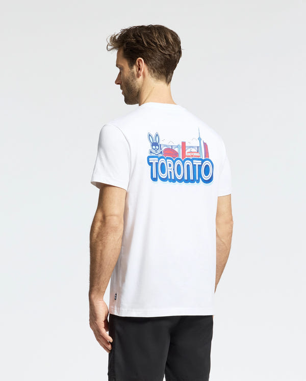 A man faces away, wearing the Psycho Bunny MENS TORONTO TEE (B6U383F200) in white Pima cotton, with a colorful hometown hero graphic of city landmarks and a cartoon rabbit on the back; one hand is by his side.