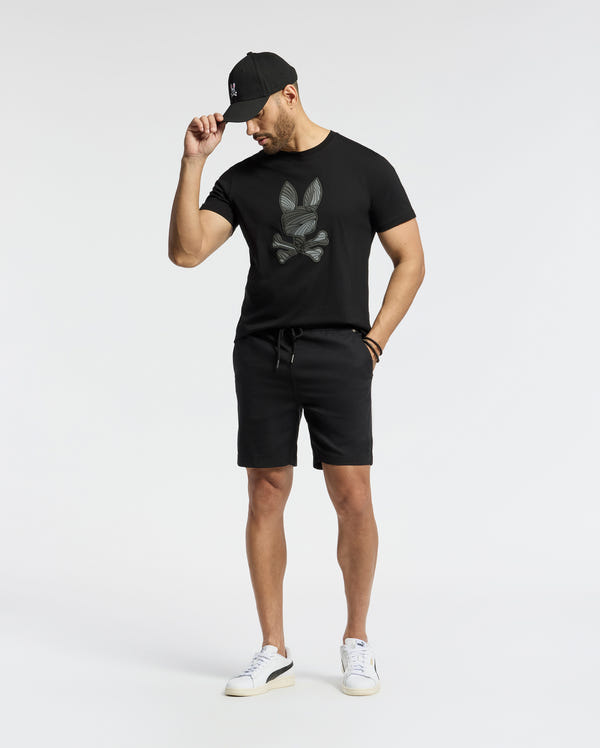 A man in a black cap, black shorts, and white sneakers wears the Psycho Bunny MENS BAXTER GRAPHIC TEE - B6U409F200. He stands against a plain white background, tilting his head down and holding his cap's brim.
