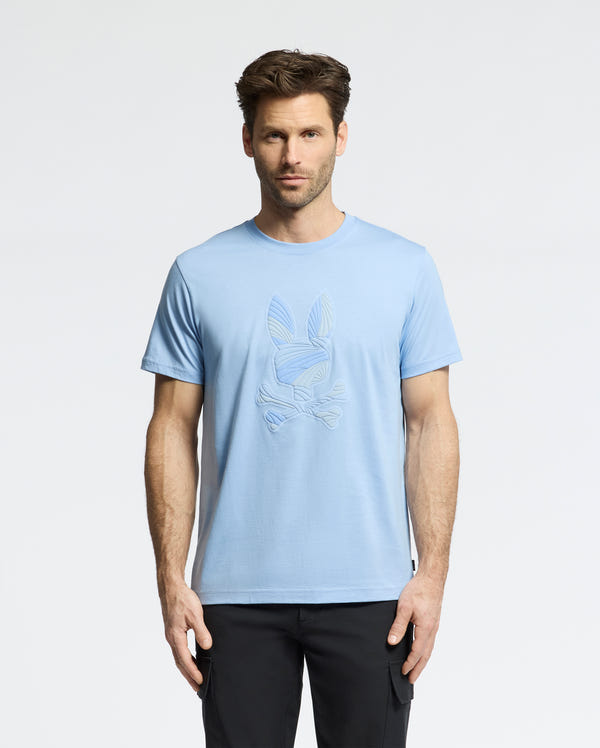 A man with short brown hair and a beard stands facing forward, wearing the Psycho Bunny MENS BAXTER GRAPHIC TEE (B6U409F200), a light blue Pima cotton shirt with a subtle bunny and crossbones design, paired with black pants against a plain white background.