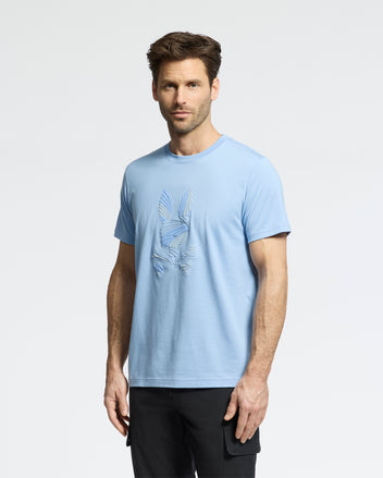 A man with short brown hair and a trimmed beard wears the Psycho Bunny MENS BAXTER GRAPHIC TEE (B6U409F200), a light blue tee with a textured bunny skull print, standing slightly angled against a plain white background.