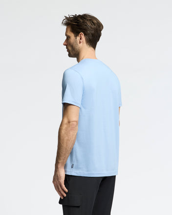 A man faces away from the camera, wearing a light blue Psycho Bunny MENS BAXTER GRAPHIC TEE (B6U409F200) and black pants, set against a plain white background.