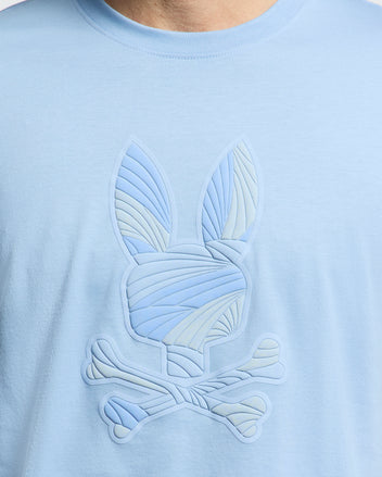 A close-up of the Psycho Bunny MENS BAXTER GRAPHIC TEE (B6U409F200) in light blue, featuring an embossed stylized bunny head with crossbones and curved line patterns.