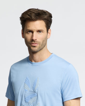 A man with short brown hair and light stubble wears the Psycho Bunny MENS BAXTER GRAPHIC TEE (B6U409F200), a light blue Pima cotton T-shirt with a subtle bunny print, standing against a plain white background.
