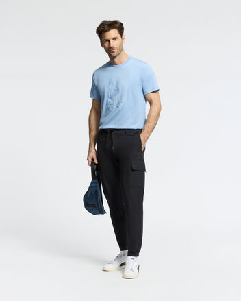 A man wearing the Psycho Bunny MENS BAXTER GRAPHIC TEE (B6U409F200) in light blue stands against a white background, paired with black cargo pants and white sneakers. He holds a small blue bag and faces the camera with a slight smile.