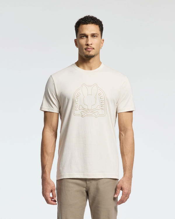 A man is wearing the MENS MELBOURNE GRAPHIC TEE by Psycho Bunny, crafted from luxurious Peruvian Pima cotton, with a stylized bunny and crossbones design on the front, against a plain white background.