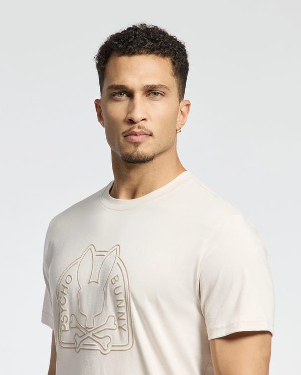 A man with short curly hair wears the eco-friendly "Mens Melbourne Graphic Tee - B6U410F200" by Psycho Bunny, made from premium Peruvian Pima cotton. It features a raised bunny and crossbones design with "Psycho Bunny" printed. He also sports earrings, all set against a light background.
