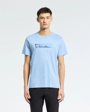 A person wearing the Psycho Bunny MENS BERNI EMBROIDERED WORDMARK GRAPHIC TEE - B6U417F200 in light blue stands against a white backdrop, perfectly pairing it with black pants. Made in Peru, the Pima cotton tee features "Psycho Bunny" text on the front.