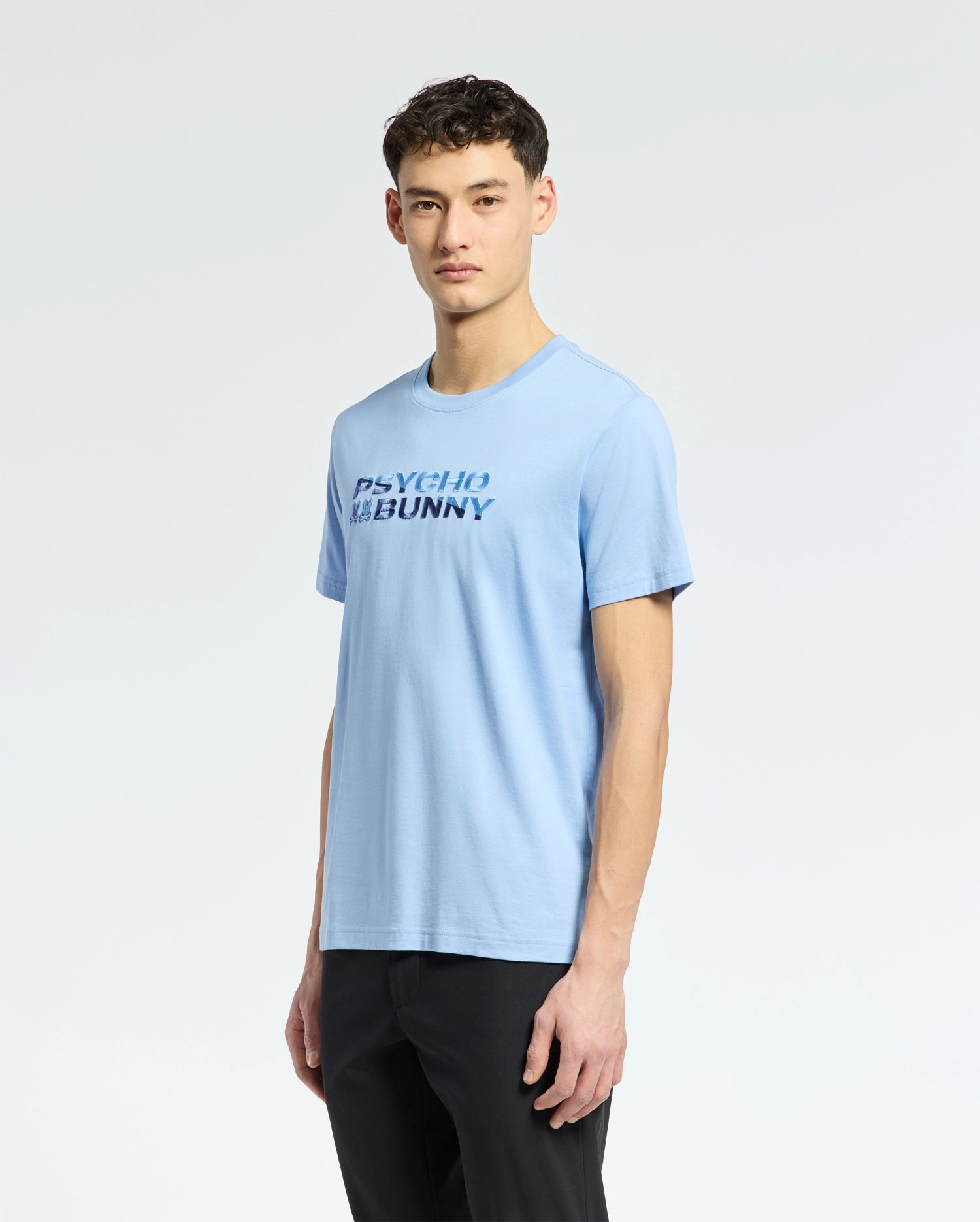 A person stands against a plain white background wearing the MENS BERNI EMBROIDERED WORDMARK GRAPHIC TEE - B6U417F200 in light blue by Psycho Bunny, paired with black pants. They have short, dark, wavy hair.