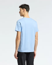 A man stands against a white background, facing away and wearing Psycho Bunny's MENS BERNI EMBROIDERED WORDMARK GRAPHIC TEE in light blue Pima cotton, paired with black pants.