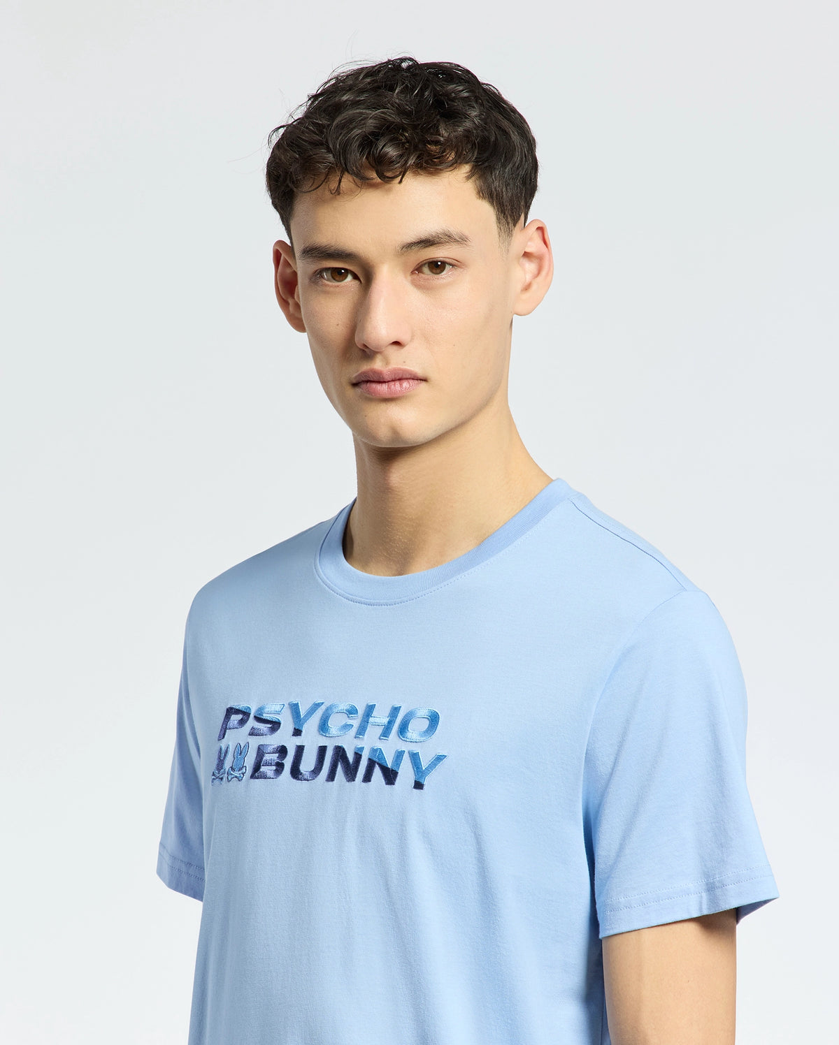 A young man with short dark hair wears the light blue MENS BERNI EMBROIDERED WORDMARK GRAPHIC TEE by Psycho Bunny, made from Pima cotton in Peru. The tee features an embroidered wordmark on the front. He stands against a plain light background, looking ahead neutrally.