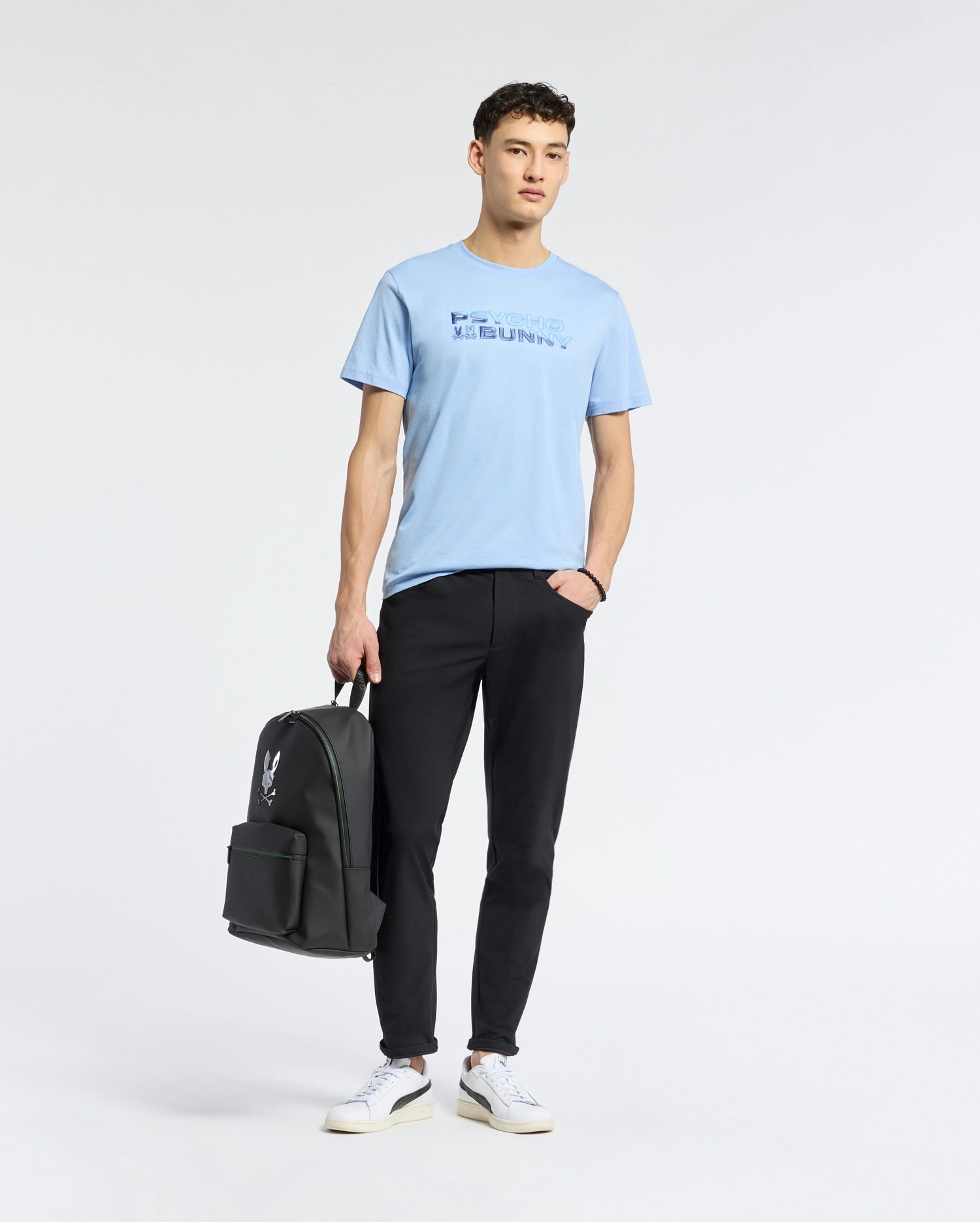 A person poses against a plain backdrop wearing Psycho Bunny's light blue MENS BERNI EMBROIDERED WORDMARK GRAPHIC TEE, black pants, and white sneakers. They hold a black backpack in one hand while the other rests in their pocket.