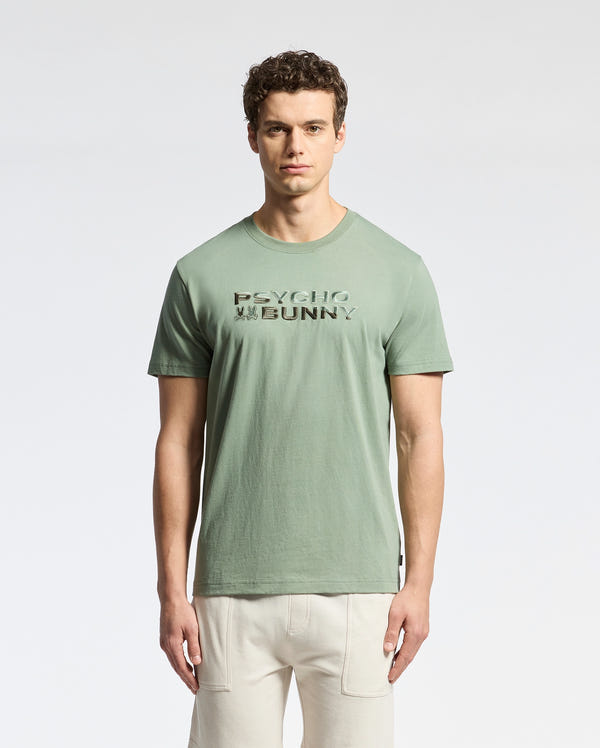 A curly-haired man wears a Psycho Bunny Berni Tee, crafted from light green Pima cotton with "PSYCHO BUNNY" text on the front and an embroidered logo, set against a plain white background.