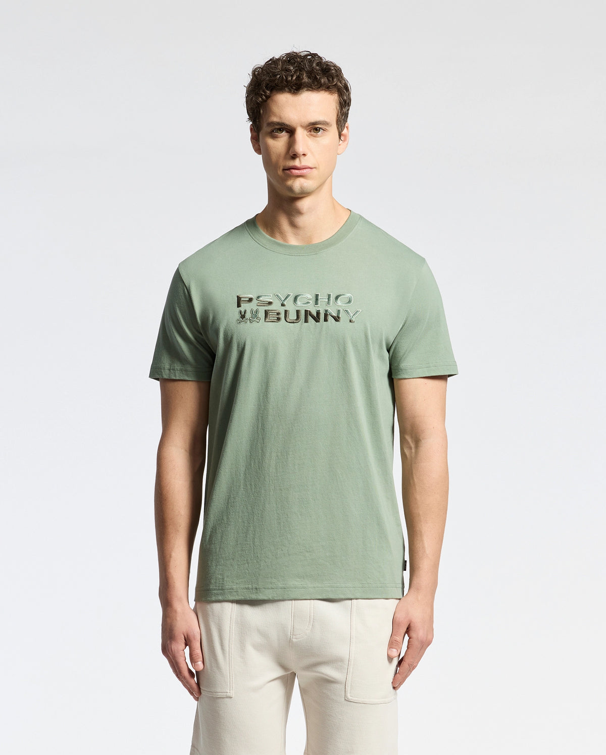 A curly-haired man wears a Psycho Bunny Berni Tee, crafted from light green Pima cotton with "PSYCHO BUNNY" text on the front and an embroidered logo, set against a plain white background.