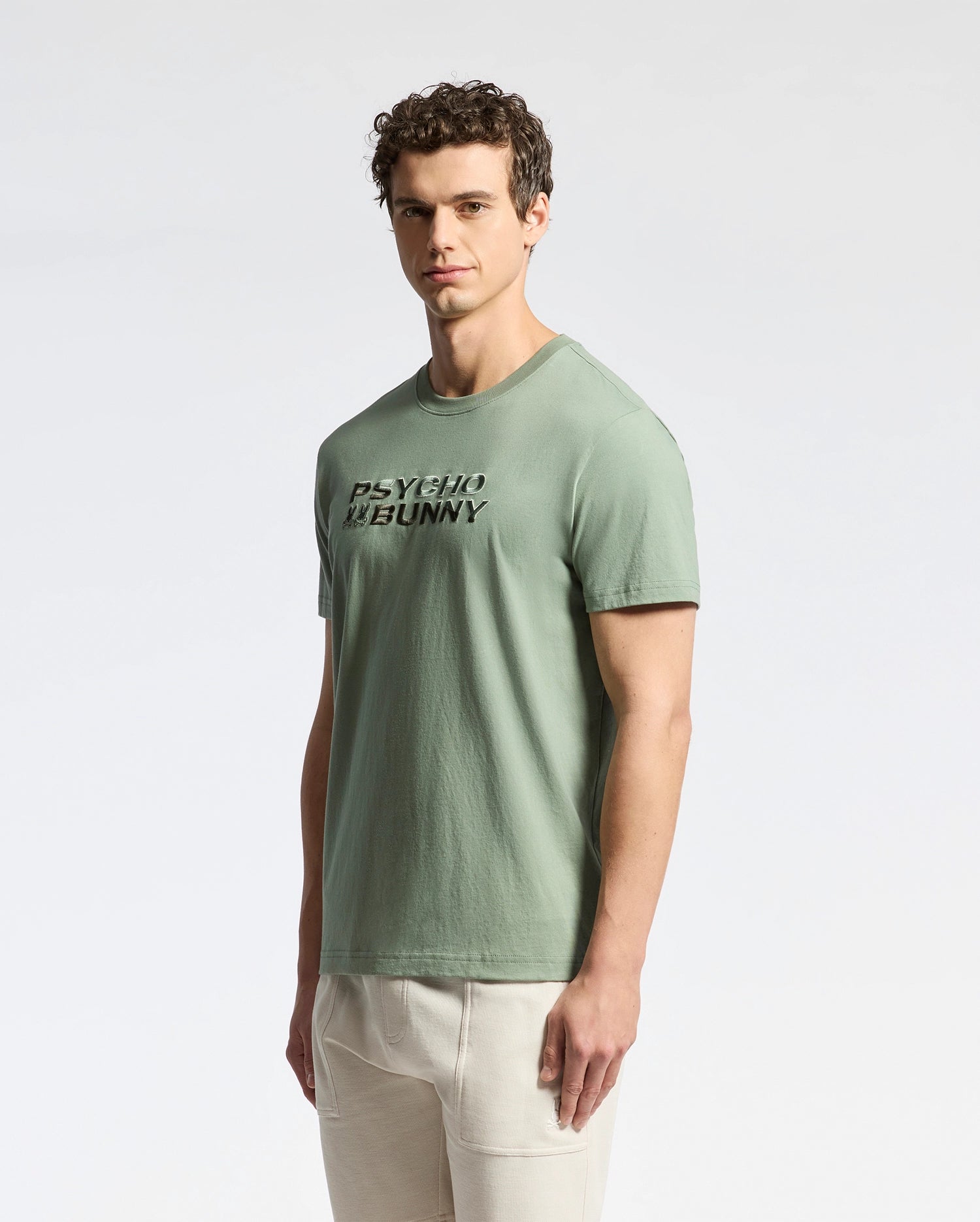 A man confidently wears the iconic Psycho Bunny MENS BERNI EMBROIDERED WORDMARK GRAPHIC TEE - B6U417F200 made of soft Pima cotton, styled with light-colored pants against a plain white background.