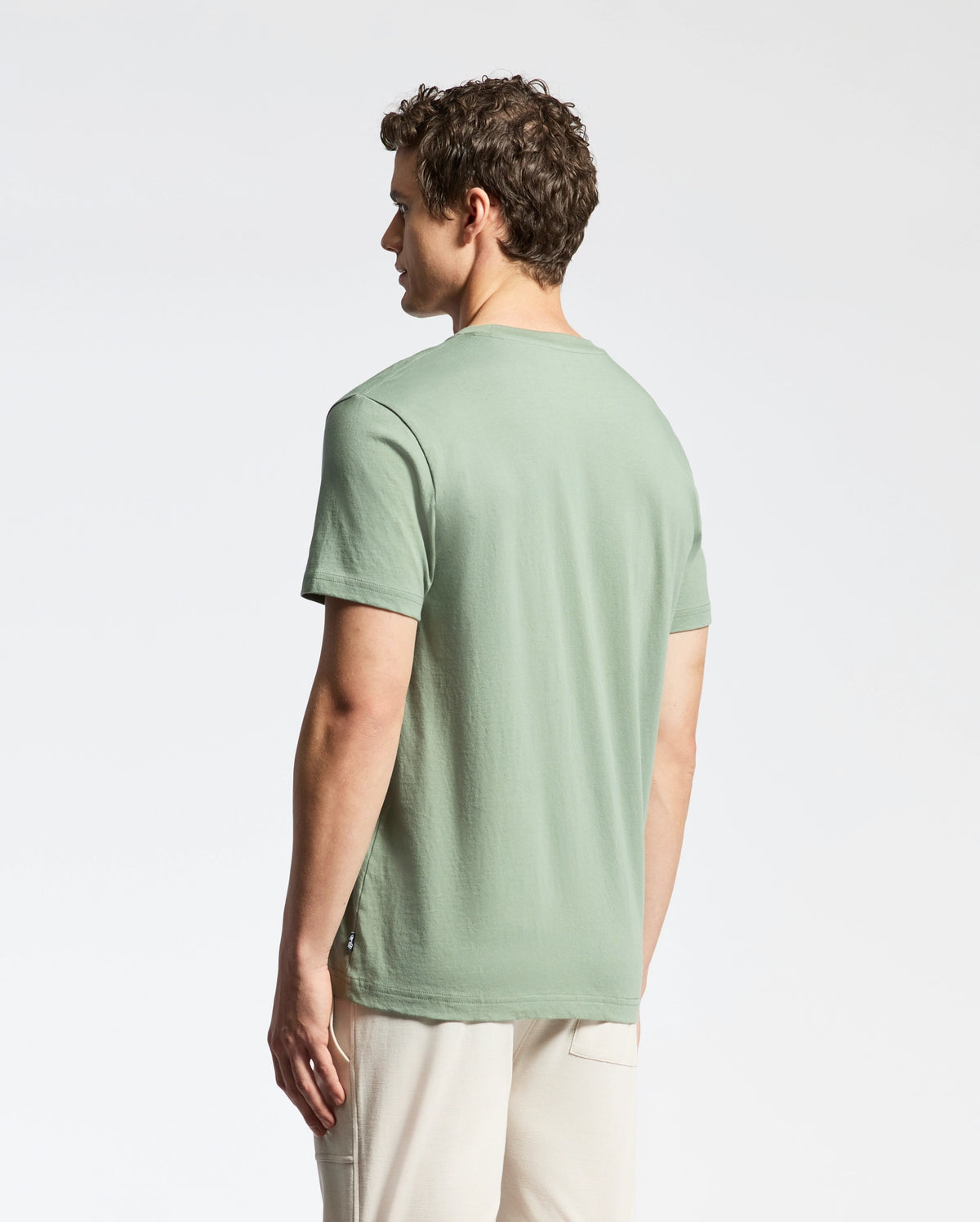 A person with curly hair stands sideways, facing left, wearing a light green Psycho Bunny MENS BERNI EMBROIDERED WORDMARK GRAPHIC TEE made from soft Pima cotton and beige pants against a plain white background.