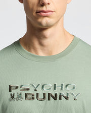 A person is wearing a light green Psycho Bunny men's Berni Embroidered Wordmark Graphic Tee (B6U417F200) made from soft Pima cotton, featuring "Psycho Bunny" text and embroidered bunny logos, with the face partially visible from the nose down.