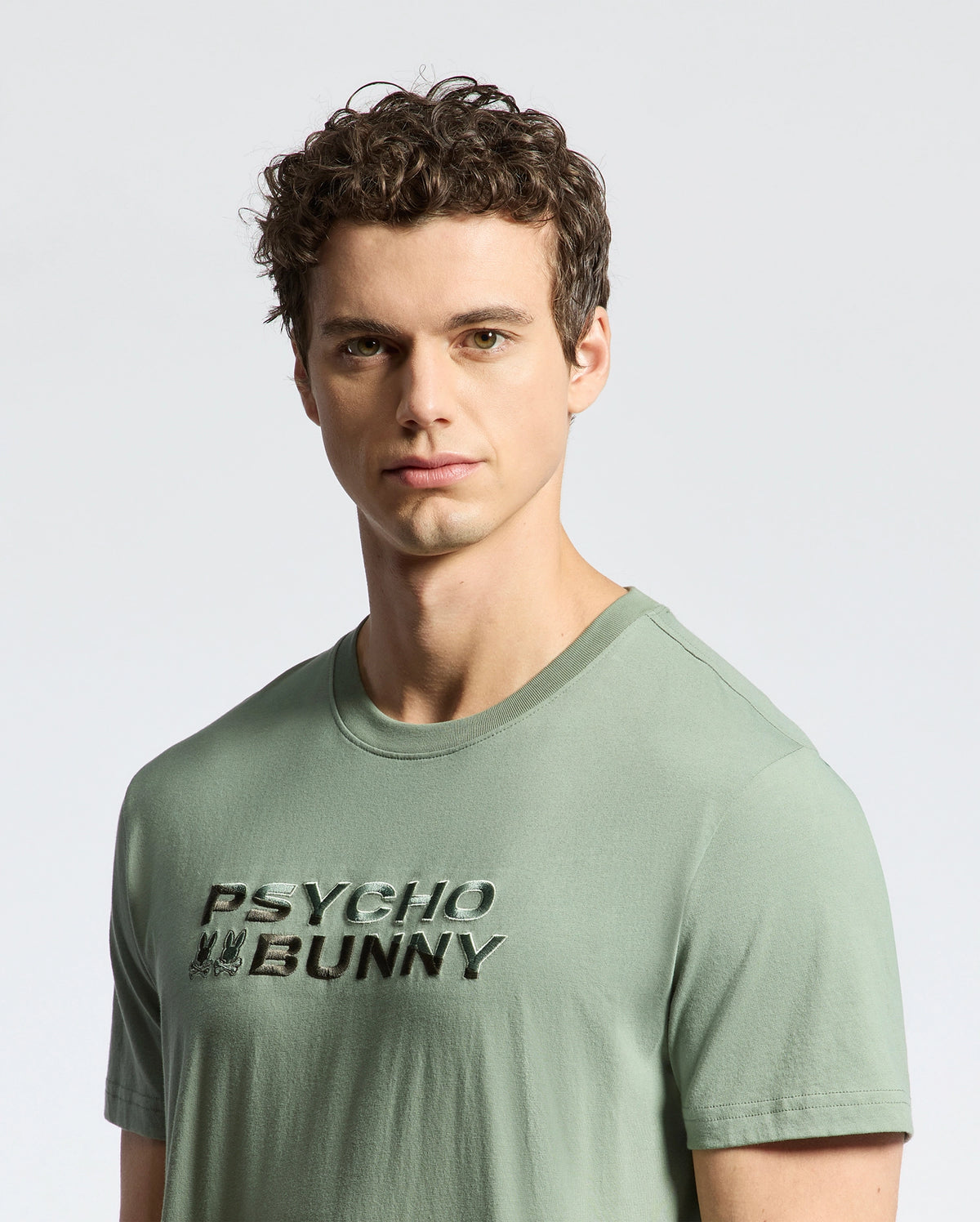 A person with curly hair dons a light green Pima cotton tee, featuring "Psycho Bunny" in bold black font. An embroidered logo enhances the Men's Berni Embroidered Wordmark Graphic Tee (B6U417F200), set against a plain light gray background.