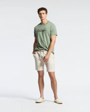 A man stands against a light grey background, wearing a Psycho Bunny men's Berni Embroidered Wordmark Graphic Tee made from soft Pima cotton. He pairs it with beige shorts and slip-on shoes, hands in pockets, complementing his short curly hair.