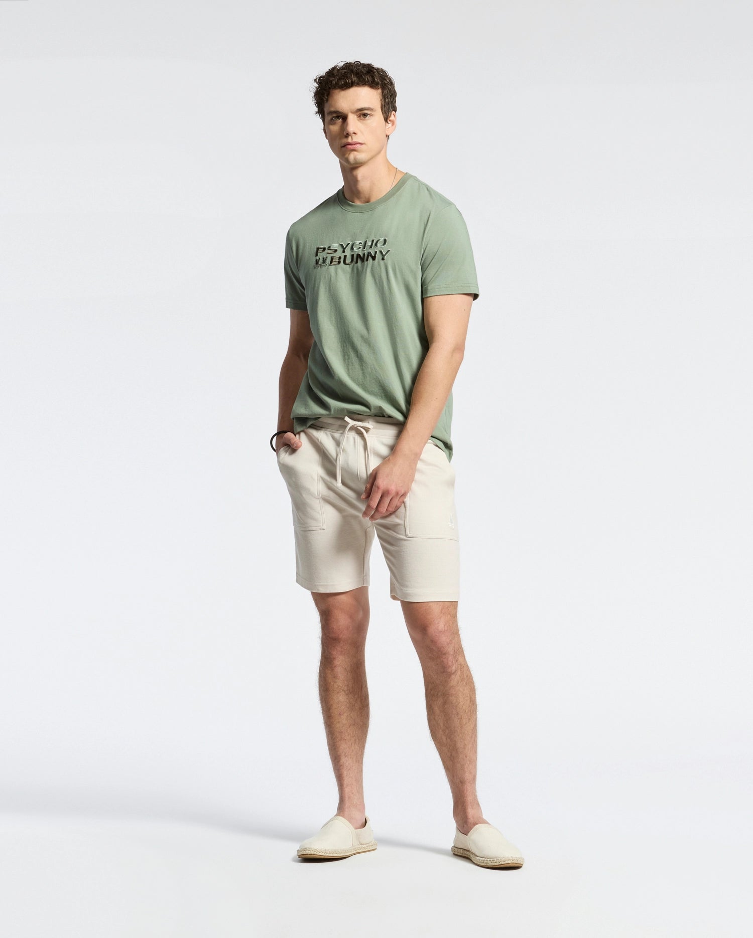 A man stands against a light grey background, wearing a Psycho Bunny men's Berni Embroidered Wordmark Graphic Tee made from soft Pima cotton. He pairs it with beige shorts and slip-on shoes, hands in pockets, complementing his short curly hair.