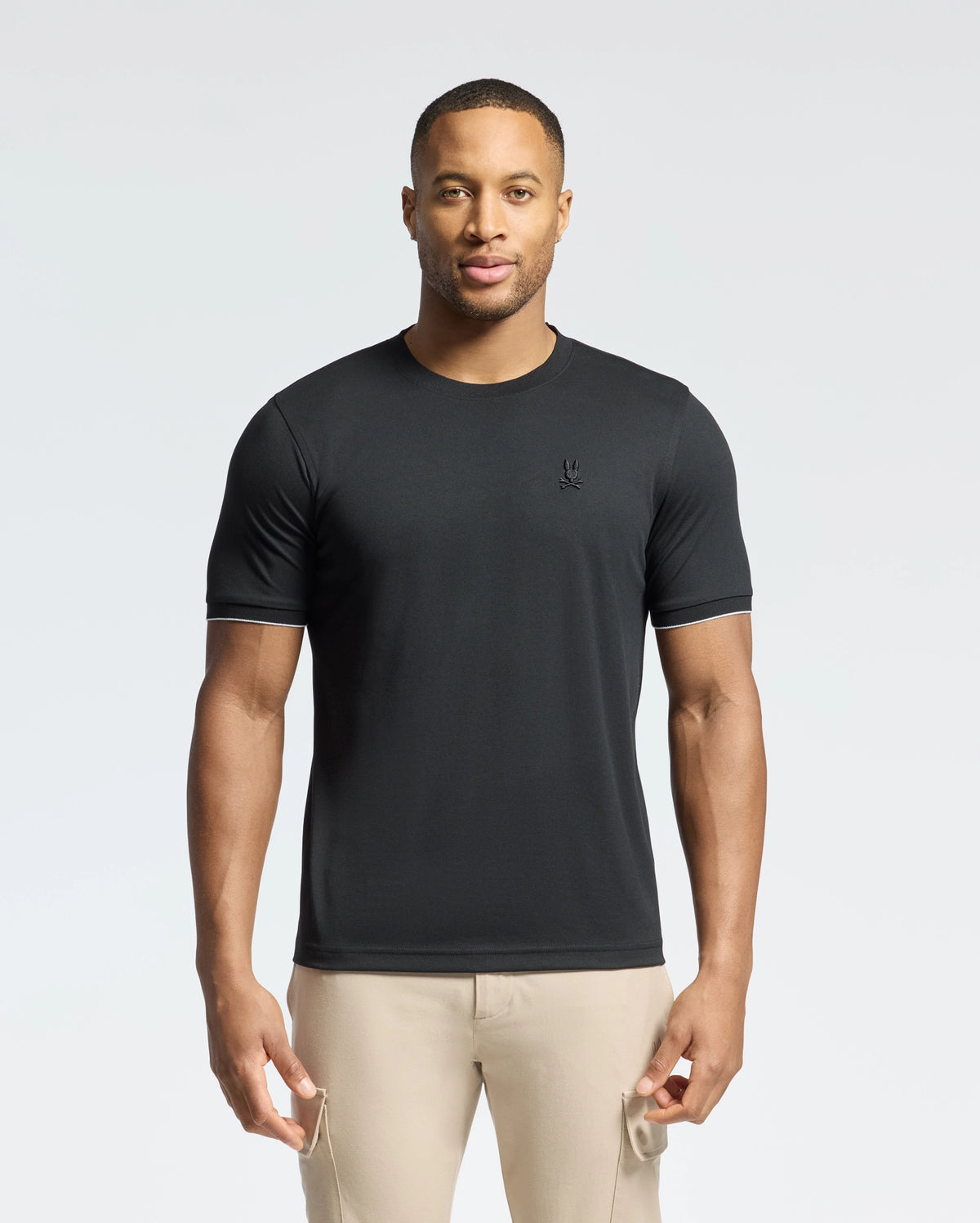In a minimalist setting, a man with short hair and a slight smile models the MENS BRYCE PERFORMANCE TEE by Psycho Bunny. Crafted from Modal-Pima Cotton in Peru, it features a small embroidered logo. Paired with beige pants, the outfit exudes effortless style and quality.